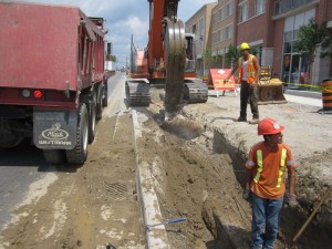 Excavator digging a trench beside a dump truck while workers observe